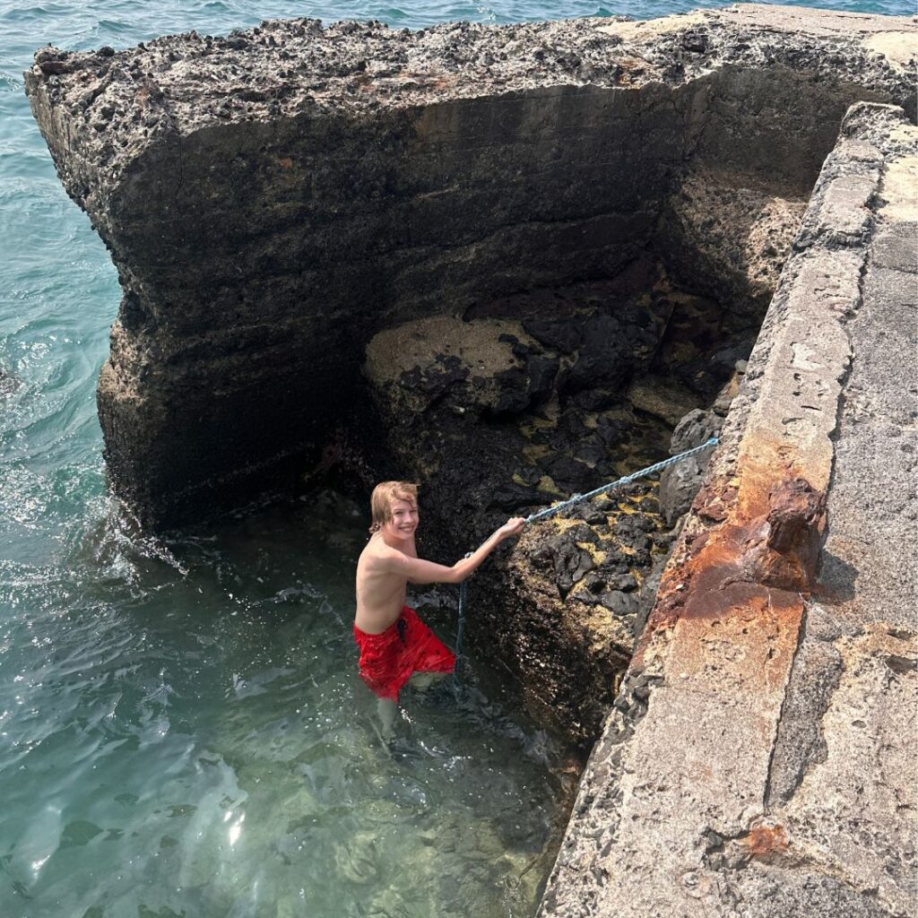 A boy with red shorts climbing out of the water onto a cliff using a rope