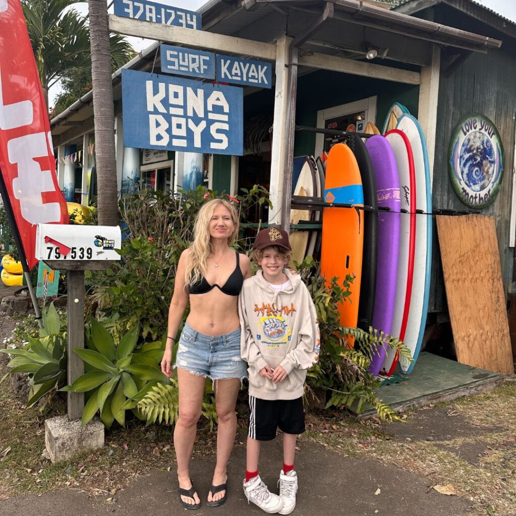 A blonde woman in a bathing suit and shorts and a boy with a hat and sweatshirt standing in front of a Kona Boys shop with surf boards