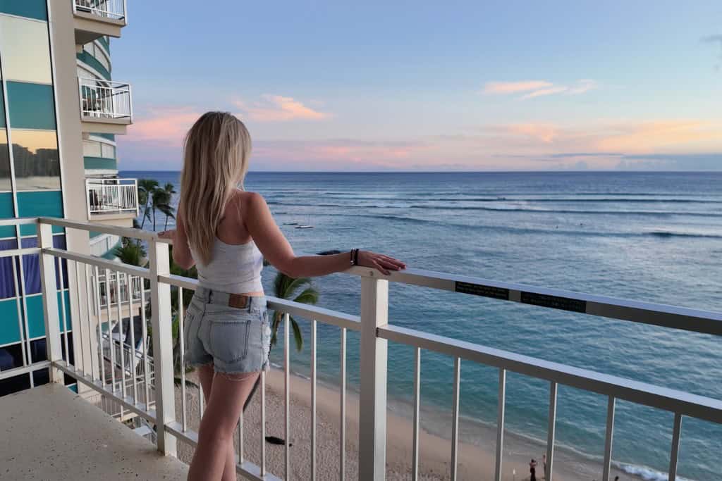 A blonde woman standing on a beachfront patio looking over the beach at sunset