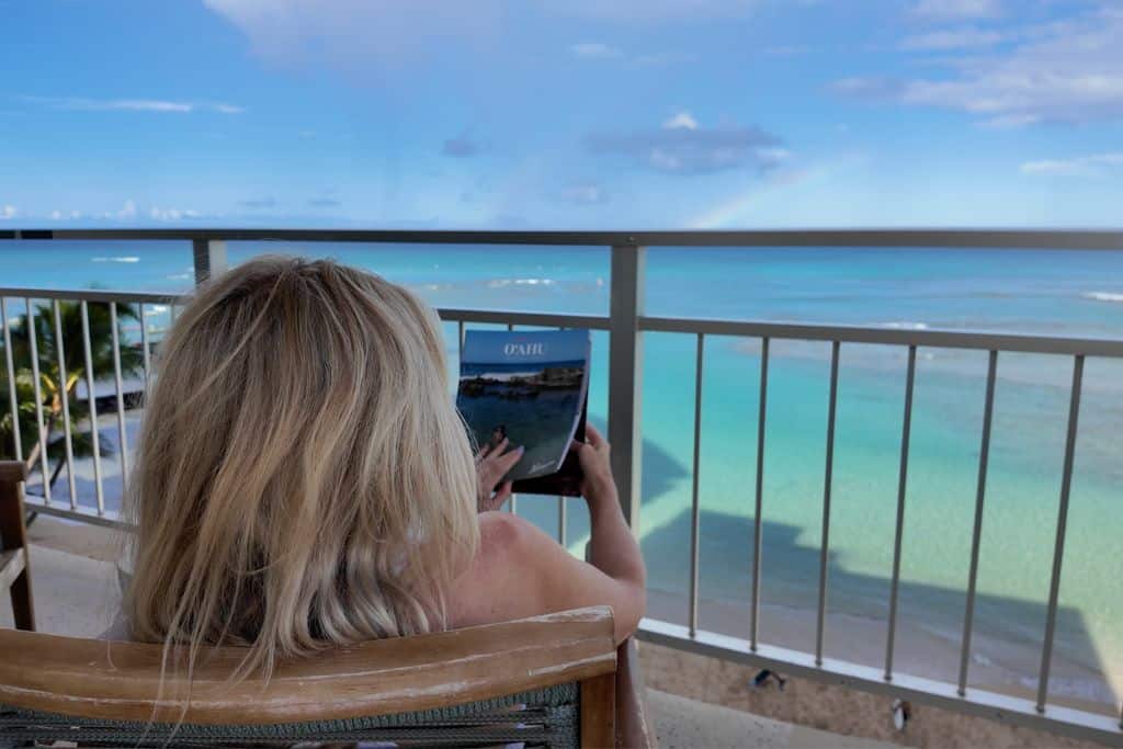 A blonde woman reading an Oahu magazine while sitting on an oceanfront patio with a rainbow out to sea