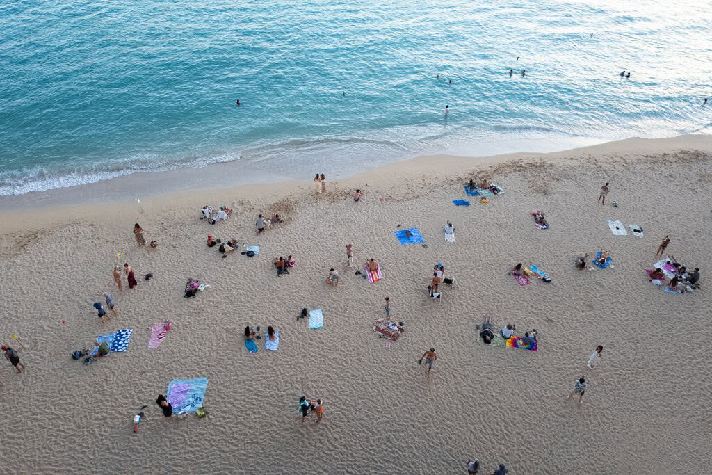 Many people sitting on a beach with colorful towels and umbrellas and clear water up ahead