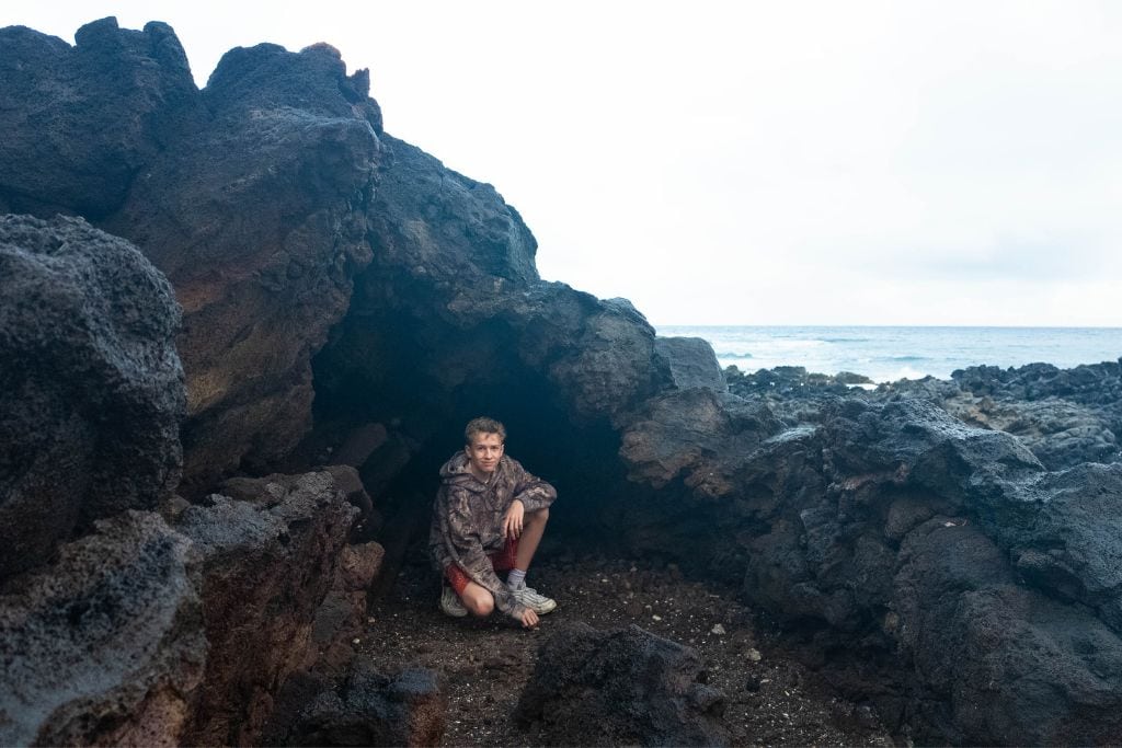 A teenage boy kneeling down surrounded by rocks with the ocean behind him.