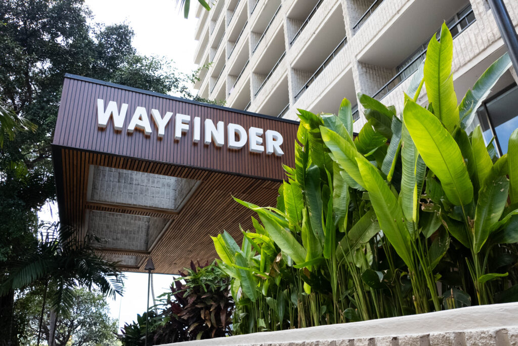 A white building with a brown sign that says Wayfinder in white, and tropical plants.