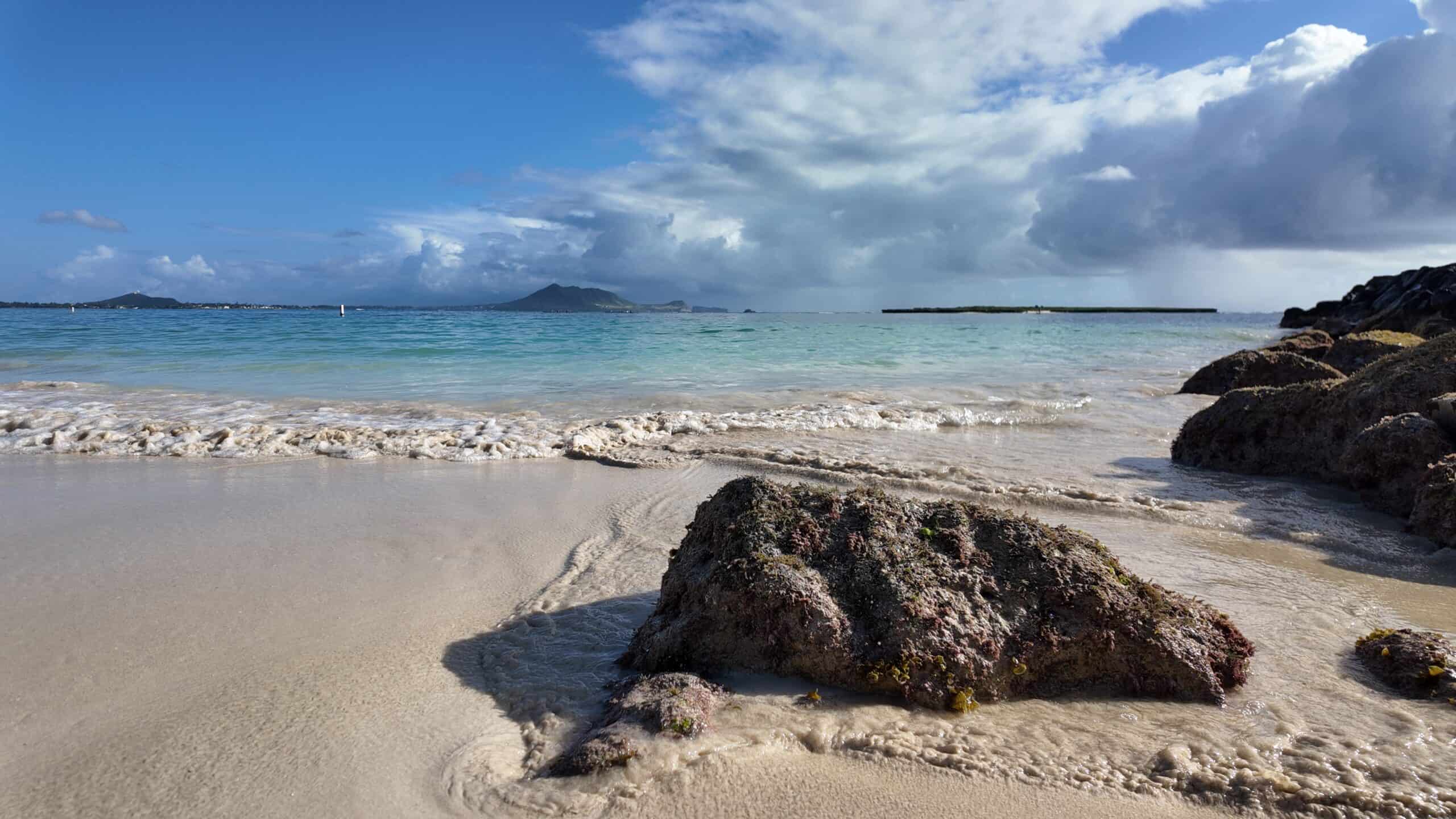 A rock in the forefront with calm turquoise ocean water and mountains in the background at Kailua Beach Park on a sunny day