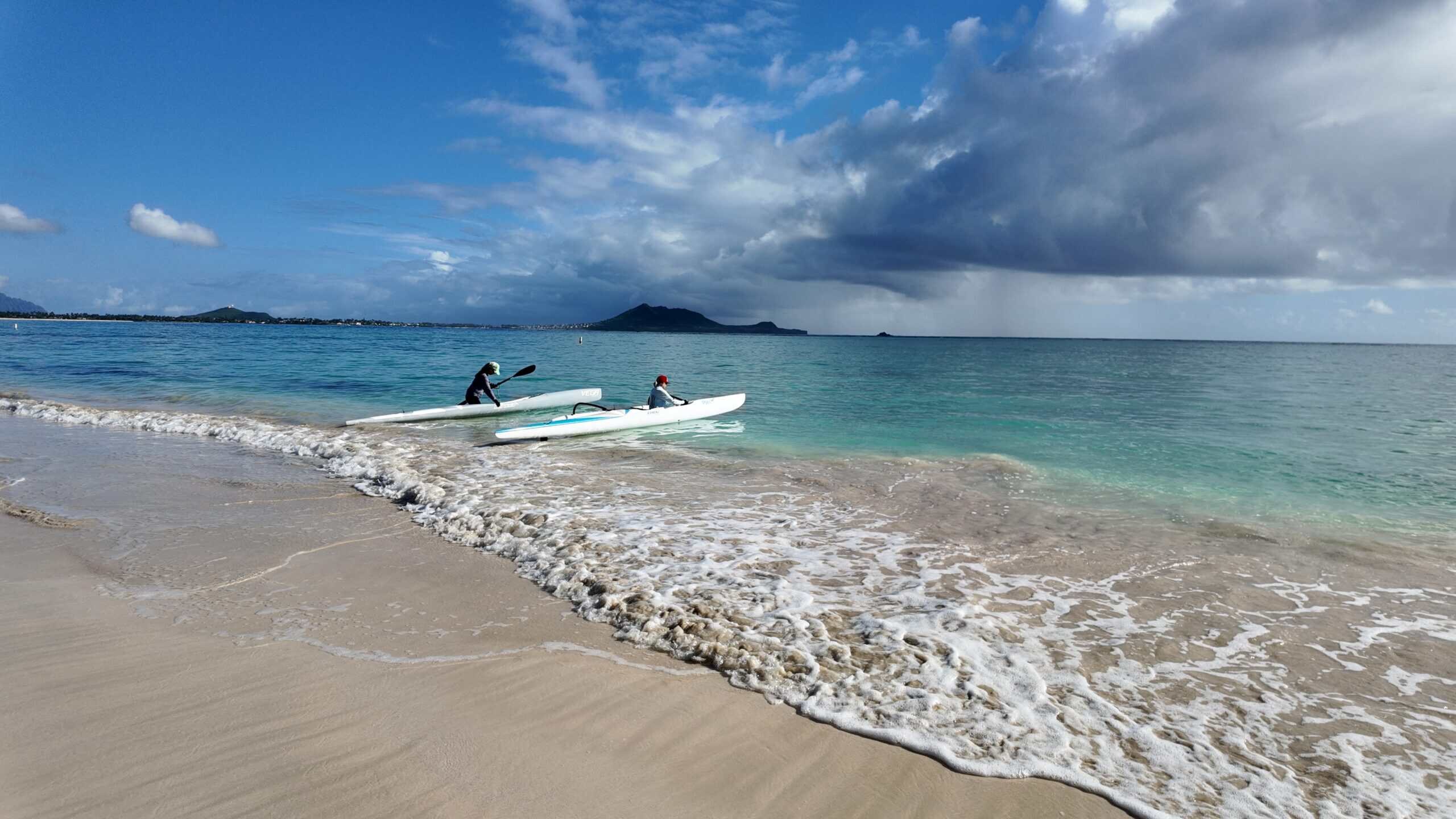 Two women getting kayaks into the clear ocean water with mountains in the background.