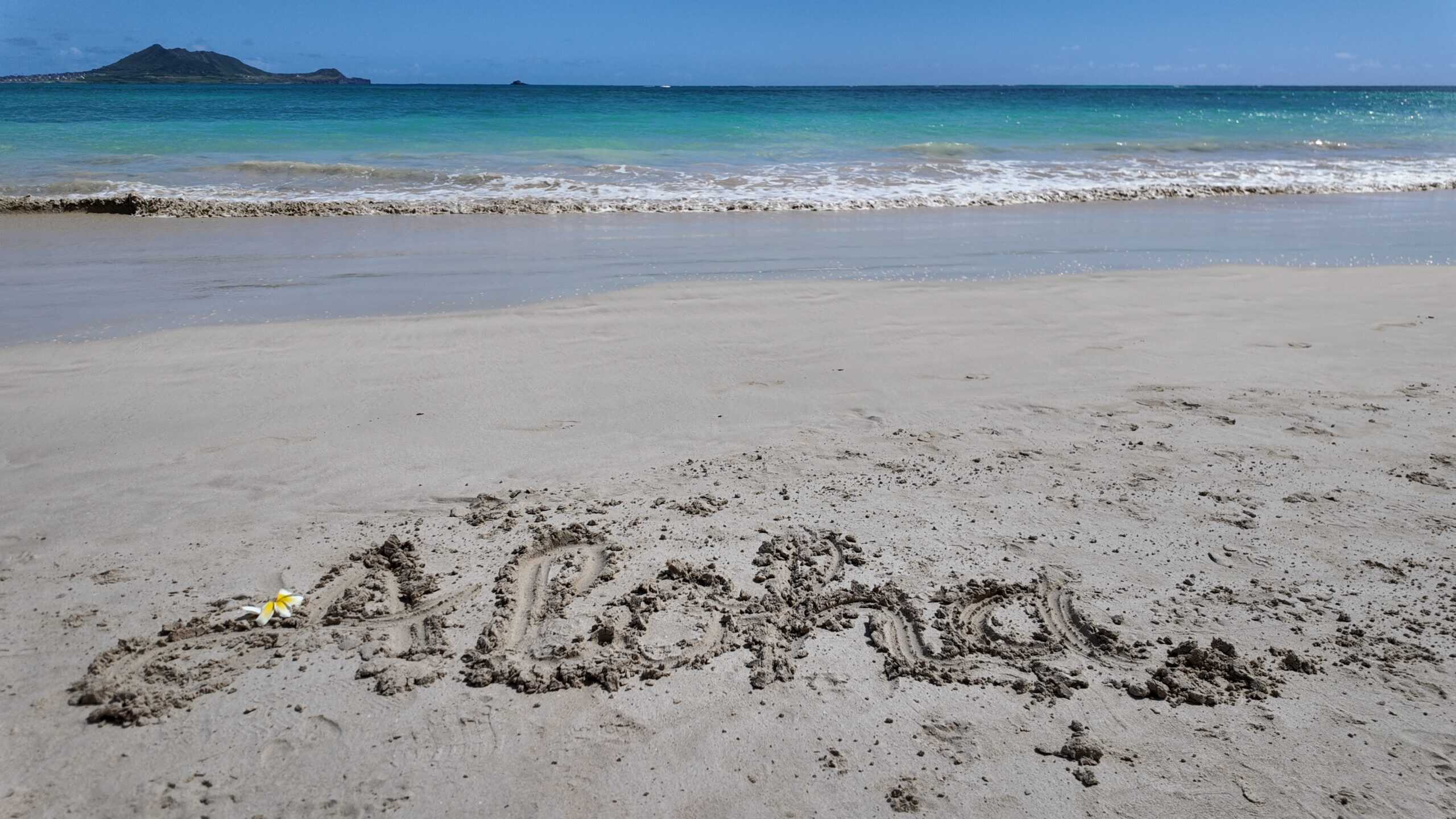 A bright white sand beach with turquoise waves and aloha written in the sand