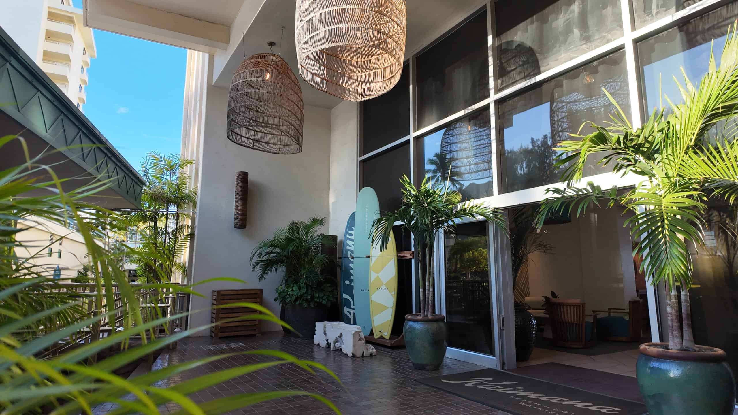 Open-air entrance to Kaimana Beach Hotel with tropical plants, surfboards, and coastal decor in Waikiki, Oahu.