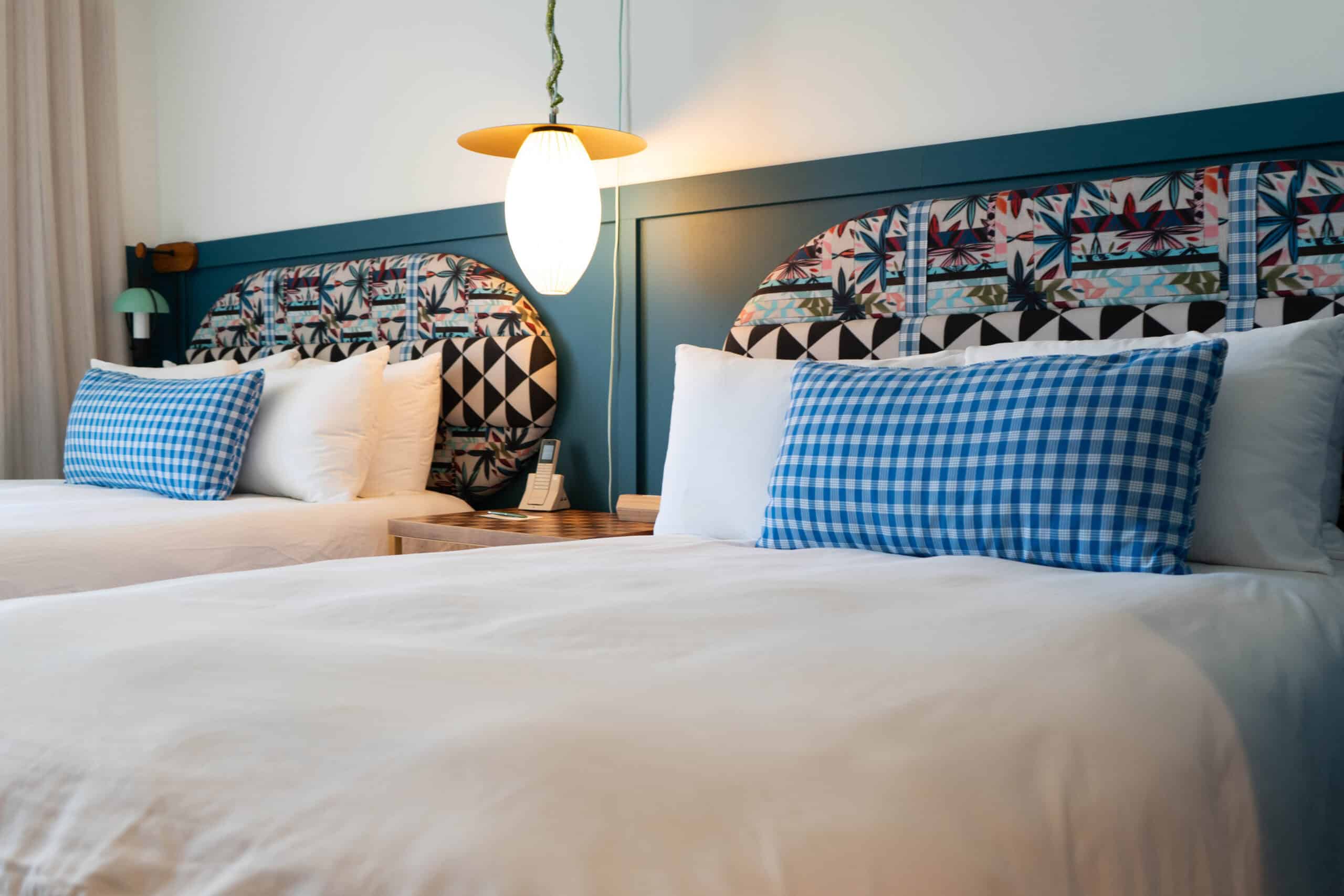 Two beds with white sheets and colorful pillows and backboards, with a light between them.