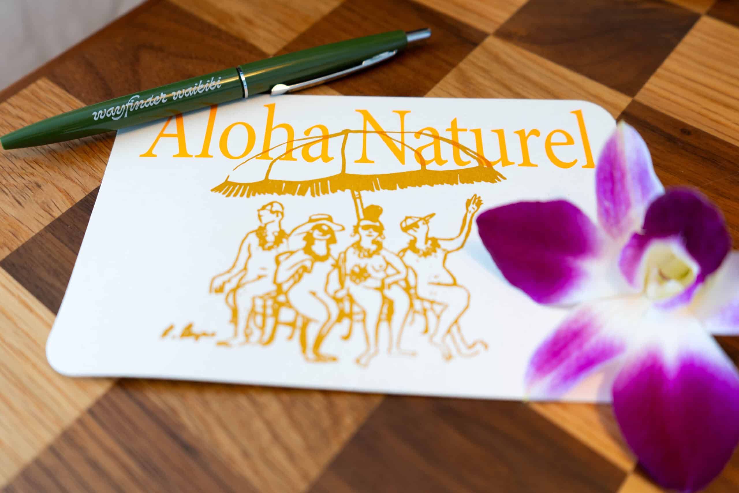 A white postcard that says Aloha Natural with sketches of people under and umbrella, a green pen that says Wayfinder Waikiki and a pink orchid