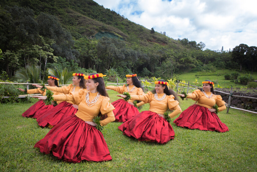 Traditional Hawaiian hula dancers in bright yellow and red costumes with mountains behind them