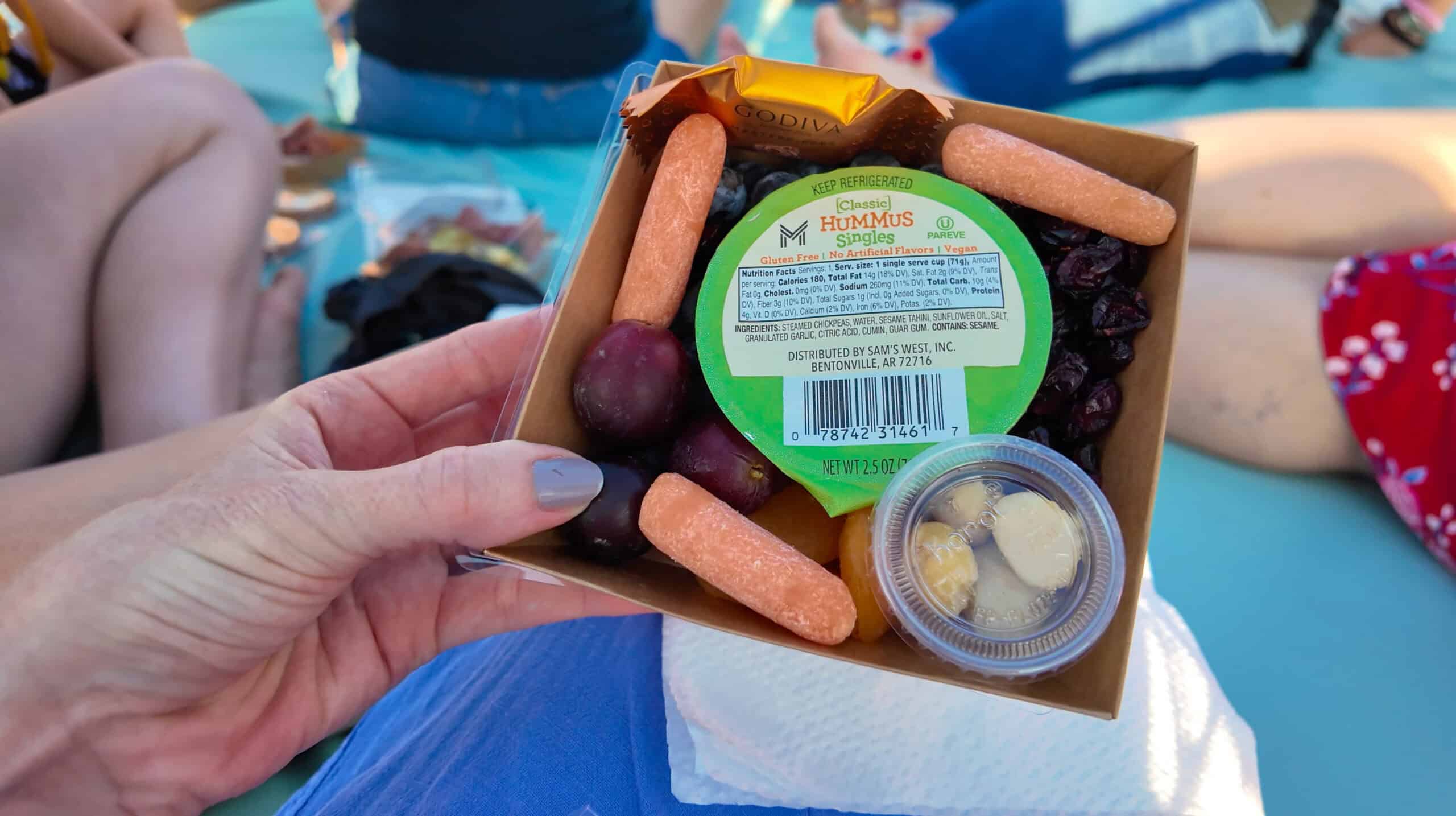 A hand with long nails holding a charcuterie board with carrots, grapes, hummus and nuts.