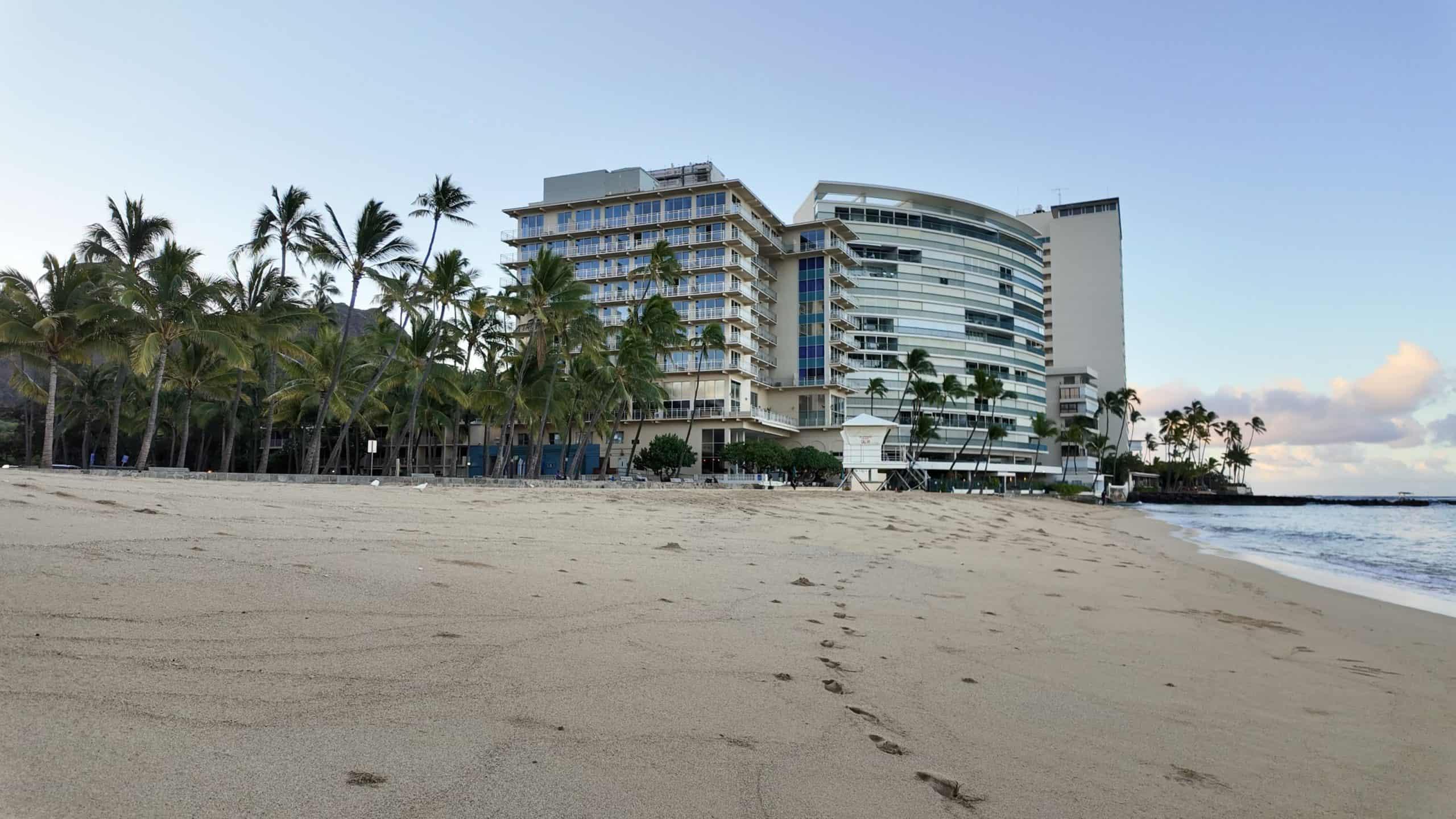 Morning view of Kaimana Beach Hotel from the sand, with footprints leading toward the oceanfront property in Waikiki.