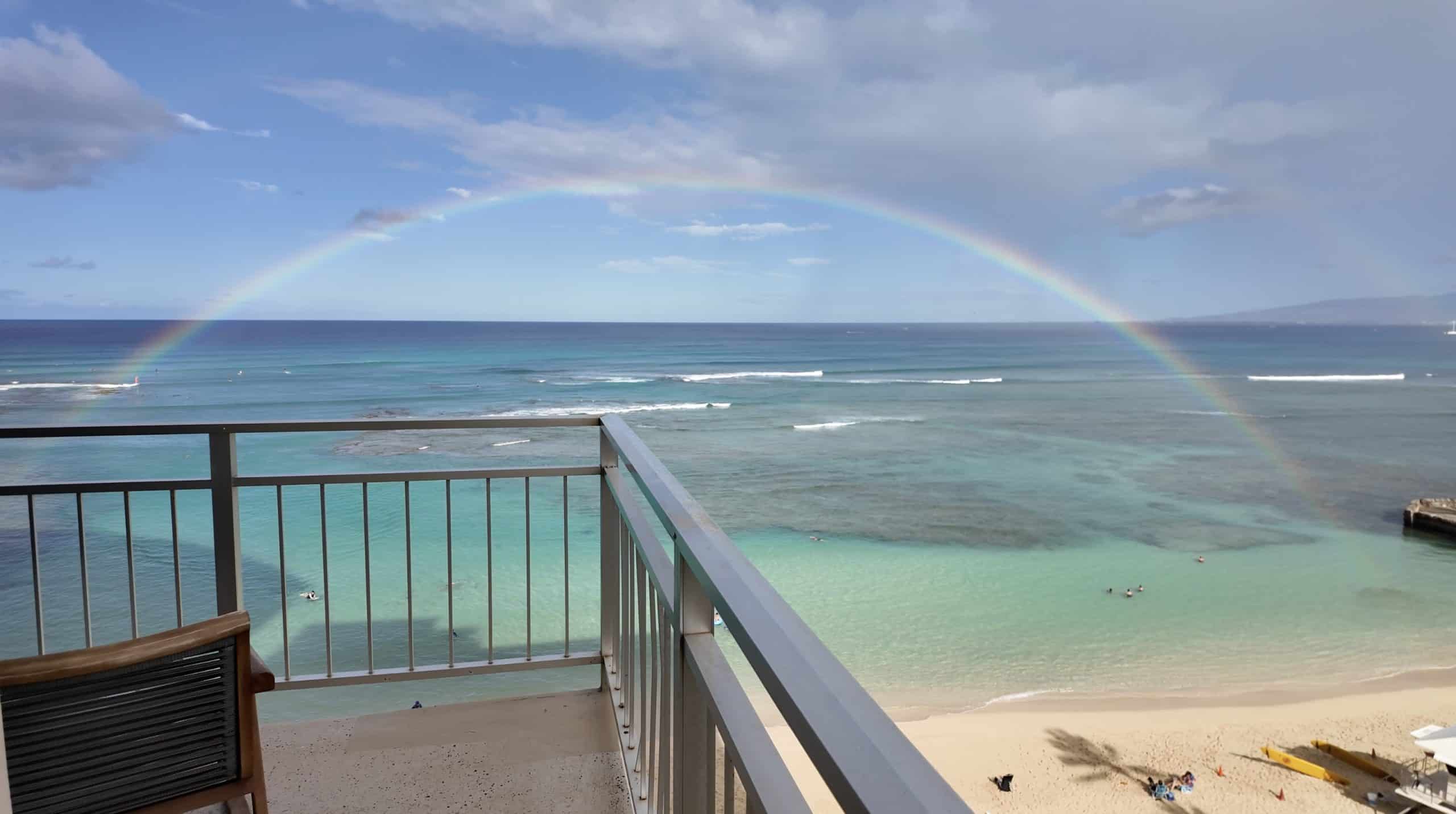 A rainbow arching across the turquoise ocean, viewed from a private hotel balcony above the sand and waves.