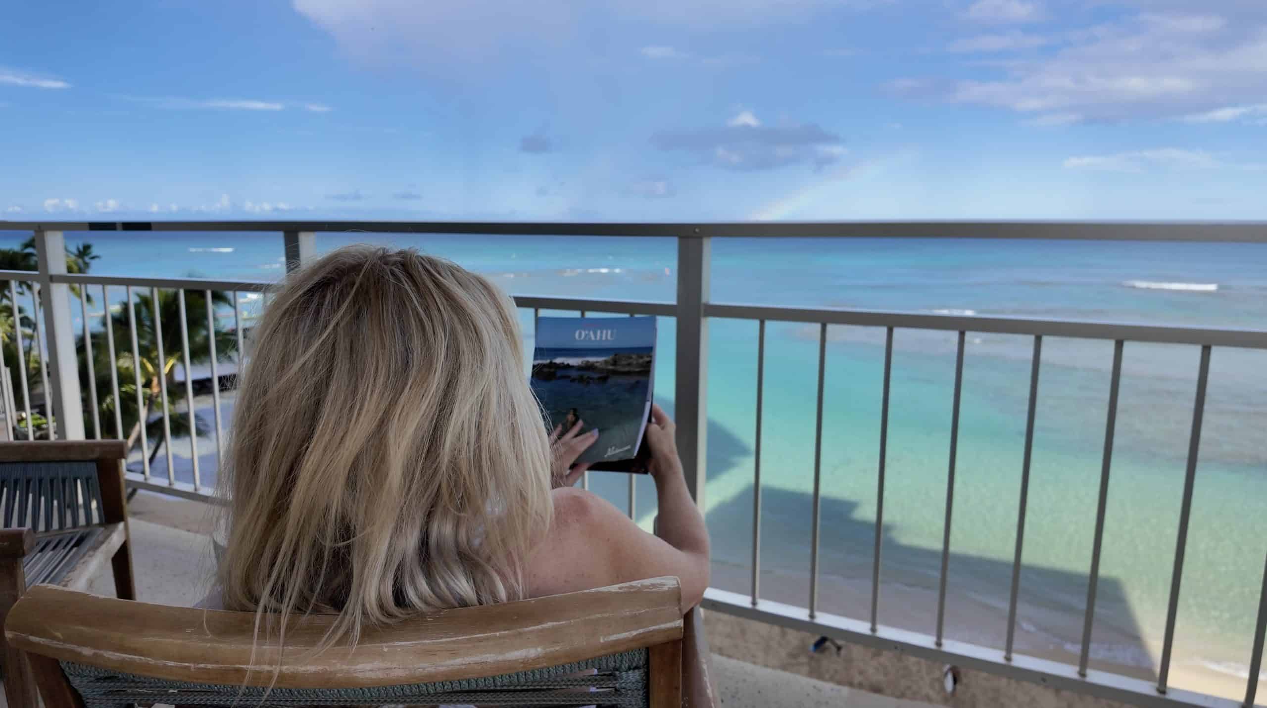 A blonde woman reading an Oahu magazine on a hotel patio looking out at a tropical ocean.